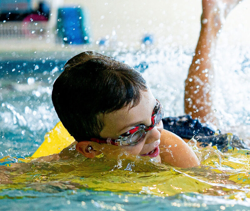 Child wearing goggles and using a kickboard during kids swim lessons, practicing strong kicking skills while having fun in a supervised indoor pool.