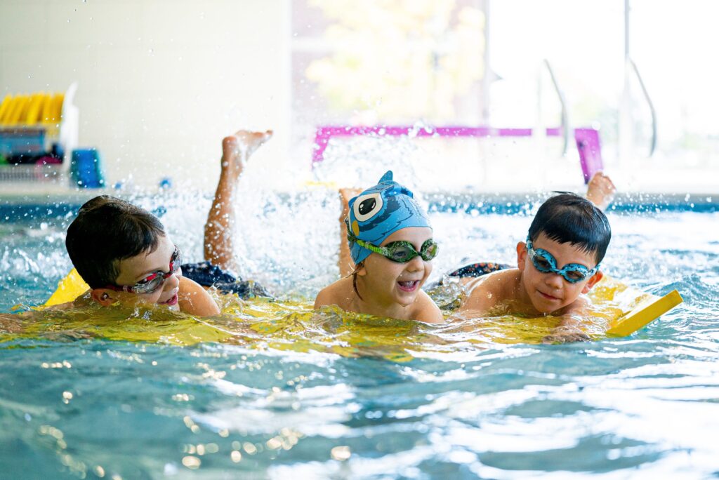 Children participating in kids swim lessons, smiling and kicking with kickboards during a fun, instructor-led swimming class in an indoor pool.